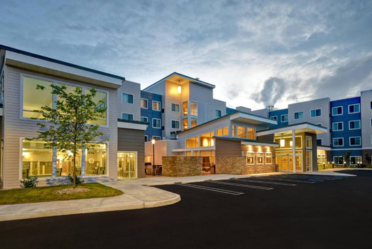 Modern three-story building with illuminated windows and wood-paneled exterior at dusk, featuring a parking lot in the foreground and a partly cloudy sky above.