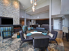 Hotel lobby with modern furniture, stone fireplace, TV, and pendant lights. Seating includes plaid and black chairs, a sofa, and round tables on a patterned carpet. Reception desk in the background.