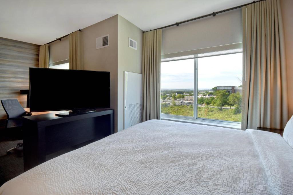 Hotel room with a bed, desk, large TV, and window overlooking a green landscape.