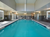Indoor swimming pool with clear blue water, surrounded by tables and chairs. Large windows let in natural light, and there are safety markings on the pool's edges.
