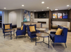Hotel lobby with blue armchairs, yellow cushions, round tables, and beige sofas. Artwork on walls and a mounted TV above the fireplace. Wood flooring and overhead lighting.