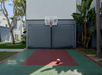 Outdoor basketball court with a hoop and net. A basketball rests on the red and green court surface, surrounded by tall trees and buildings.