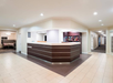 Hotel lobby with a central reception desk, tiled flooring, and a small snack area in the background.
