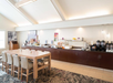 A bright dining area with a wooden table and six chairs, a long counter with breakfast items, coffee machines, and built-in lighting on the ceiling.