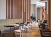Modern restaurant interior with wooden accents, a row of tables and chairs, and a green patterned carpet. Beige and brown tones dominate the space.