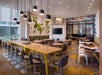 Modern cafe with long tables, chairs, and hanging lights. A green abstract wall design is visible, along with a large TV. White and brown seating areas are organized around the space.