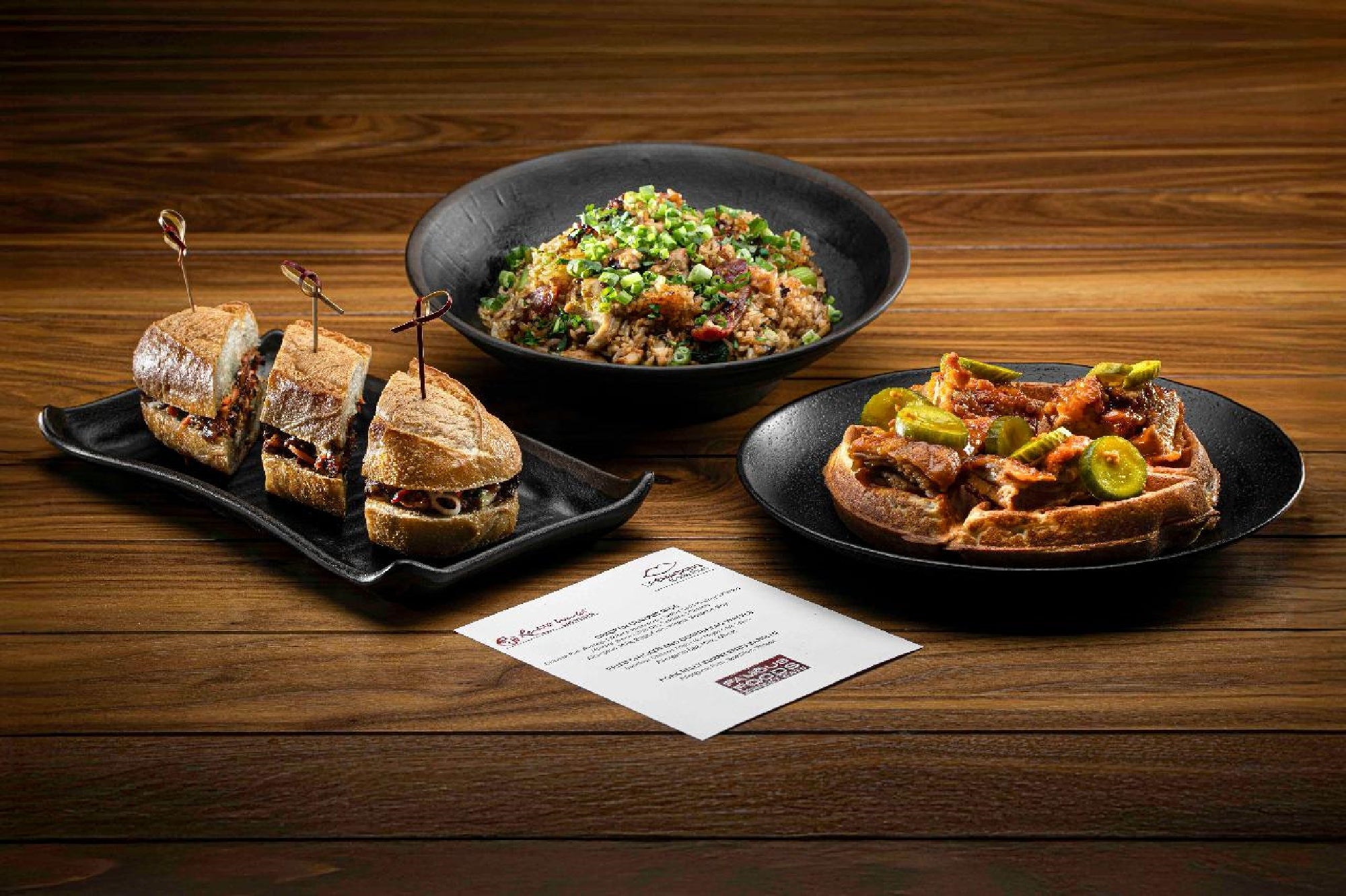 Three plated dishes on a wooden table: a sandwich cut in three, a bowl of grain salad with greens, and an open-faced sandwich with meat and pickles, alongside a printed menu card.