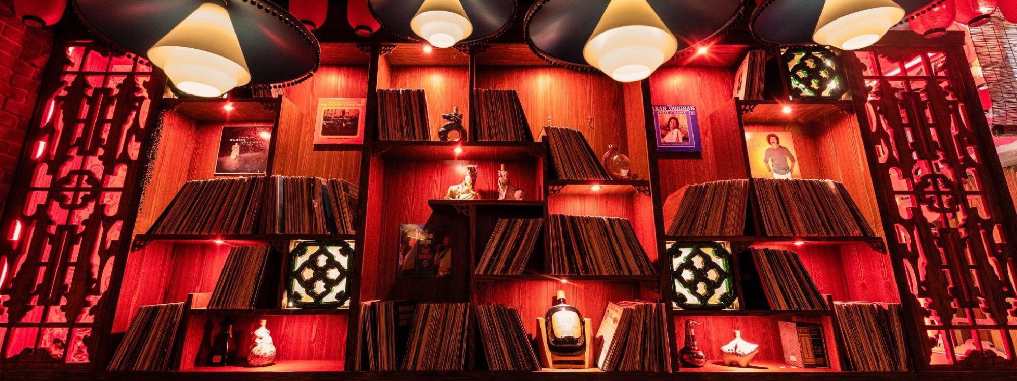 A warmly lit wooden shelf displays vinyl records, small decorative items, framed photos, and bottles, with pendant lights overhead.