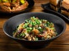A black bowl filled with fried rice, pieces of meat, and chopped green onions, placed on a wooden table with sandwiches in the background.
