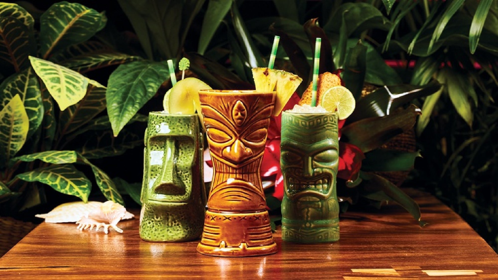 Three tiki drinks in decorative ceramic mugs with straws and fruit garnishes are displayed on a wooden table, with leafy plants in the background.