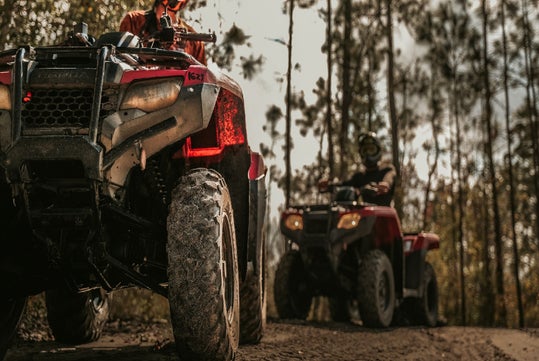 Two people riding red all-terrain vehicles on a dirt path through a forested area, viewed at ground level with one ATV in the foreground.