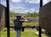 A man aims a shotgun at a clay target in an outdoor shooting range, standing between two wooden panels with trees and clouds in the background.