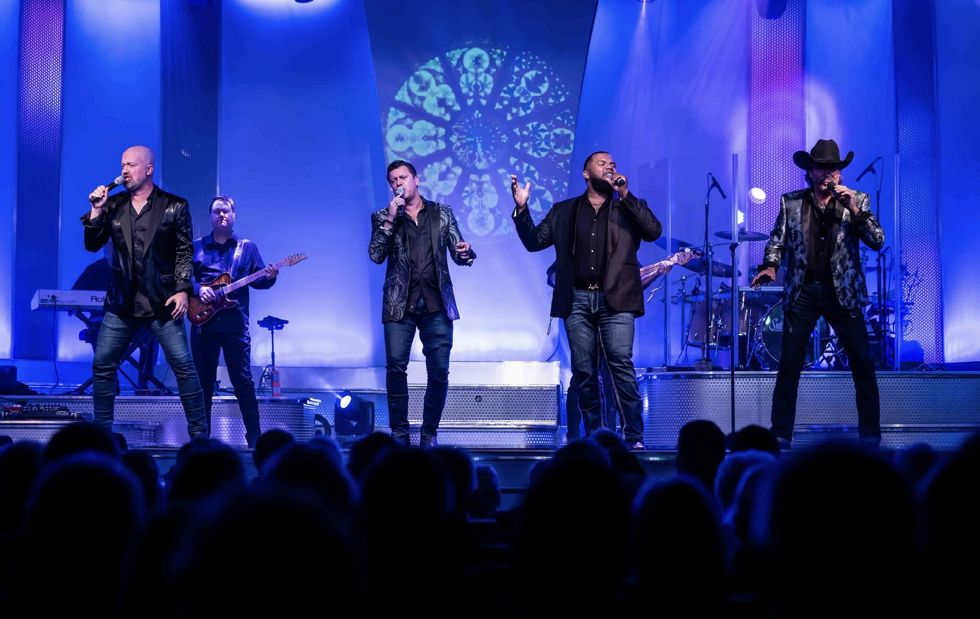 A band performs on stage under blue lighting, with four male vocalists singing at the front and musicians playing instruments in the background.
