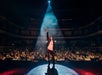 Performer stands on stage under a spotlight, facing a large audience in a theater, with one arm raised.