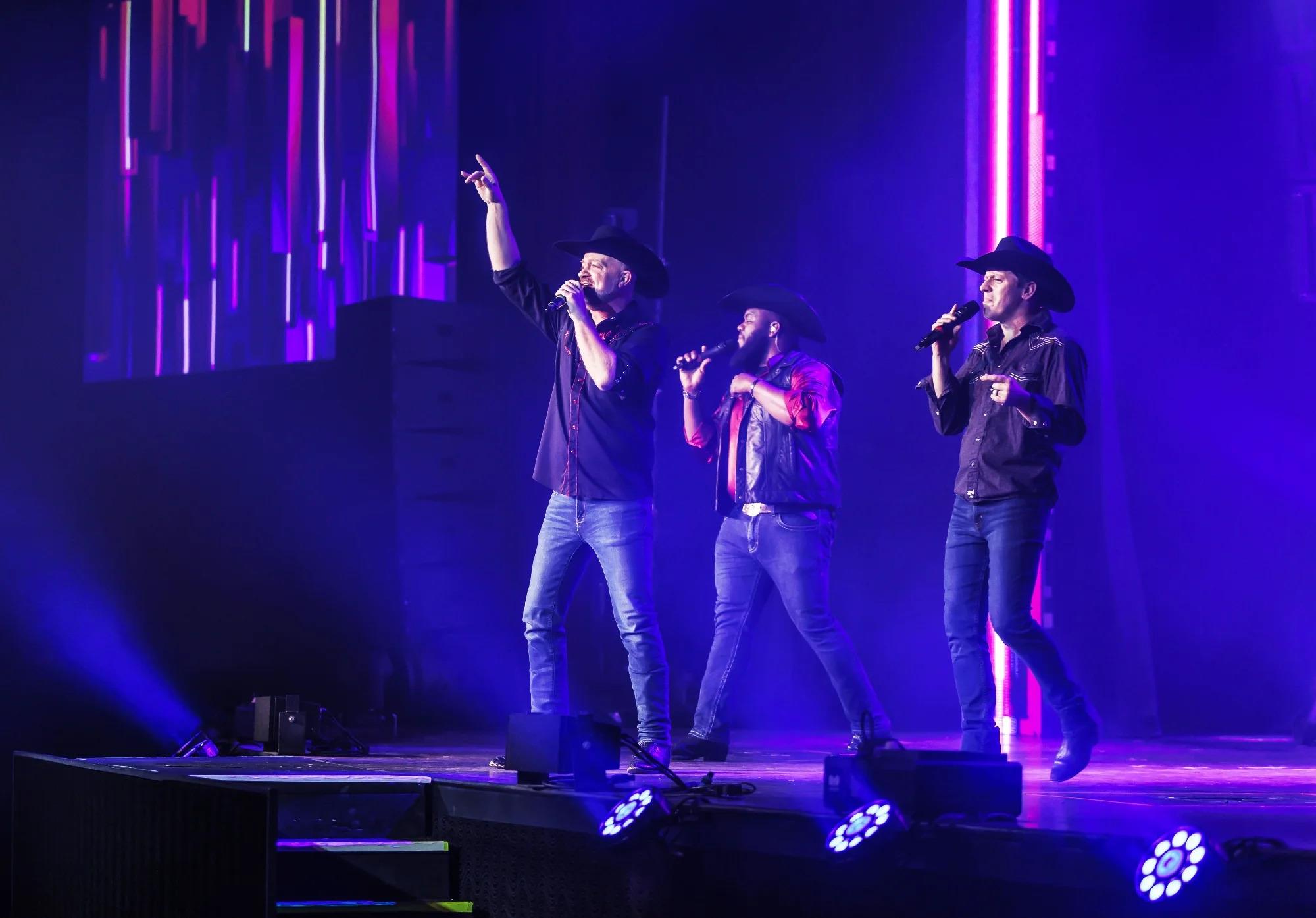 Three men in cowboy hats and casual clothes sing on stage under purple and blue lighting, holding microphones, with a colorful LED backdrop behind them.
