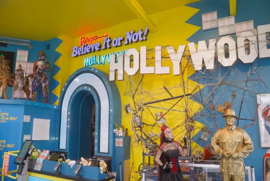 Ripley’s Believe It or Not! Hollywood entrance with colorful signs, a gold-painted performer, mannequins, and unusual displays against a bright blue and yellow wall.