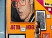 Two people look at a large portrait of Justin Bieber made from colored candy, displayed on an orange wall with other framed artworks.