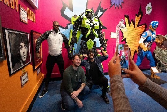 Two people crouch and two stand by a colorful robot statue inside a vibrant exhibition room. Another person takes a photo of them with a smartphone.