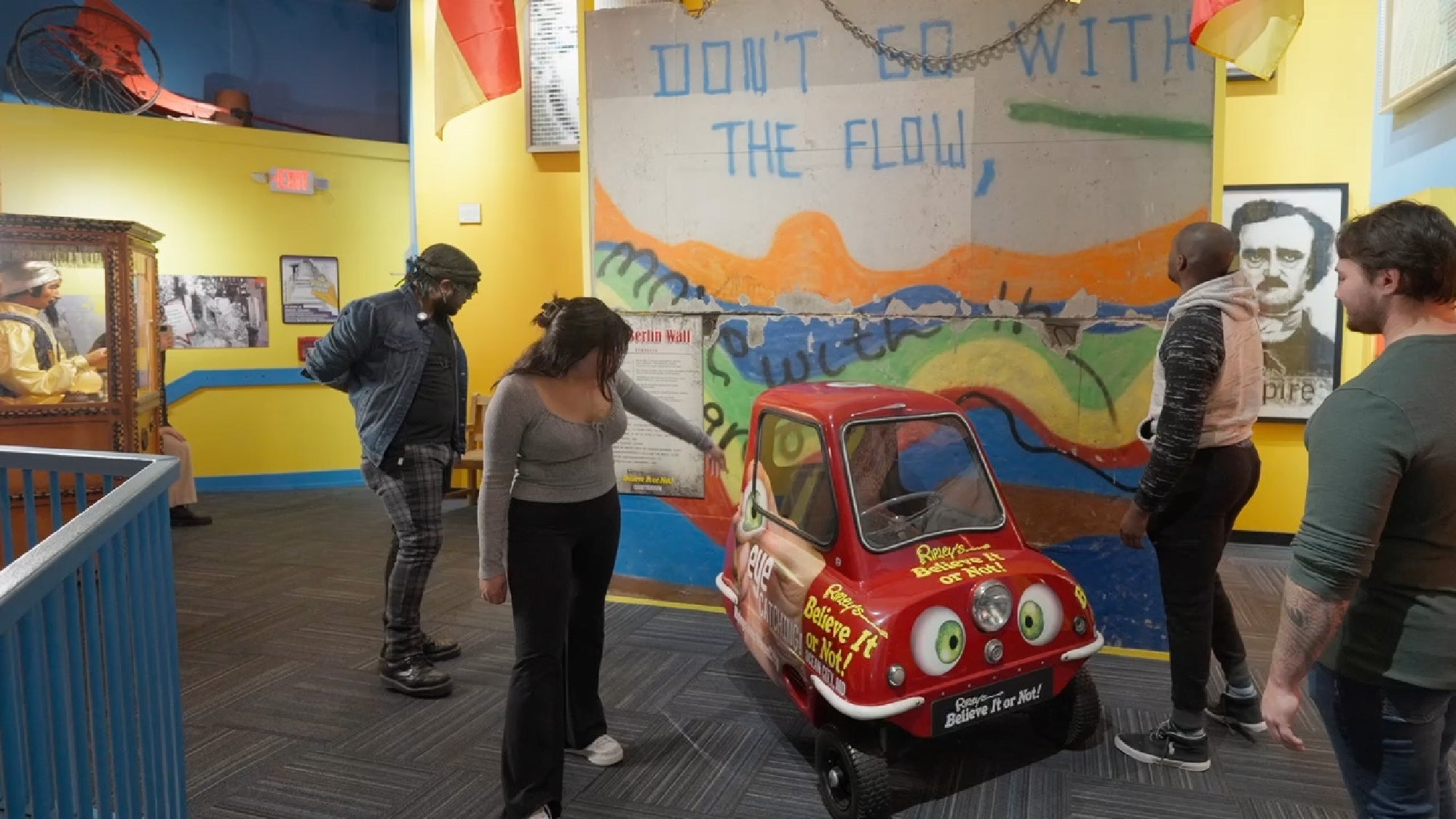 People look at a small, cartoonish red car on display in a colorful room with various artworks and signs.