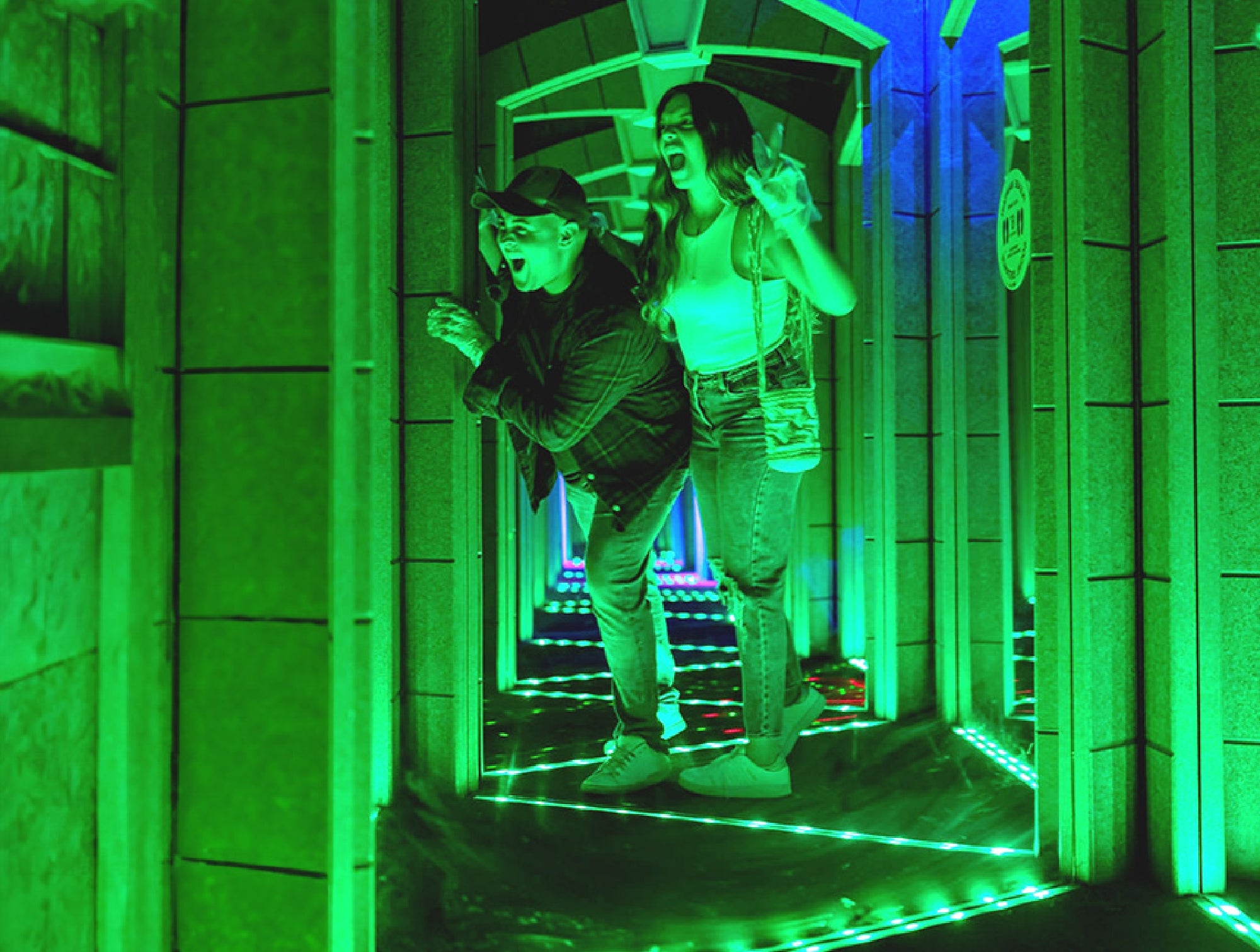 A person in a cap and a person in denim shorts are startled in a green-lit mirrored maze.