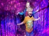 Two children walk through a tunnel with vibrant purple and pink lights, smiling and holding onto side rails.