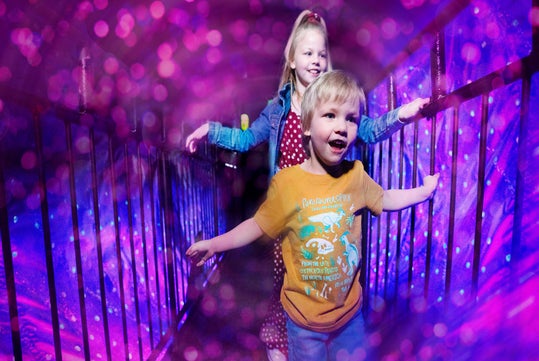 Two children walk on a colorful, illuminated bridge, surrounded by purple lights and bokeh effects.