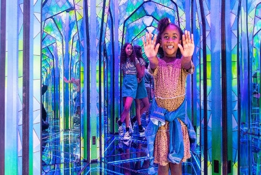 A young girl in a dress with her arms straight out in front of her with an older girl standing behind her in the Mirror Maze in Orlando.