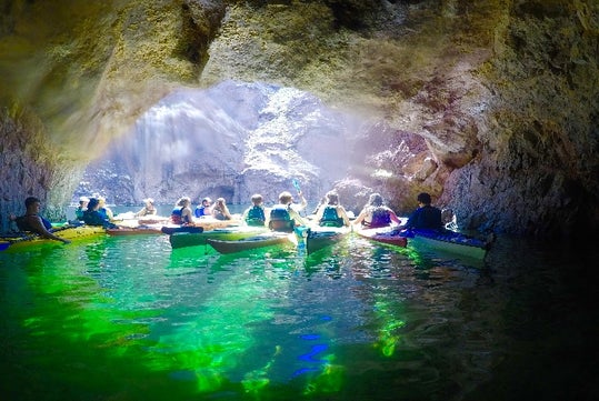 A group of kayakers sitting in the water at the mouth of a cave with the sun shining on them.