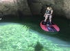 A woman standing on a red paddle board in a cave with sun shining on the water to her left.