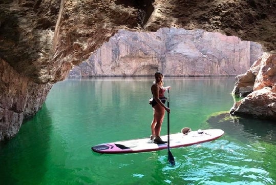 A woman standing on a paddle board at the mouth of a cave with green water under her and the sun shining in the background.