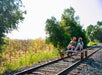 Two people wearing helmets ride a railbike on a train track surrounded by trees and greenery.