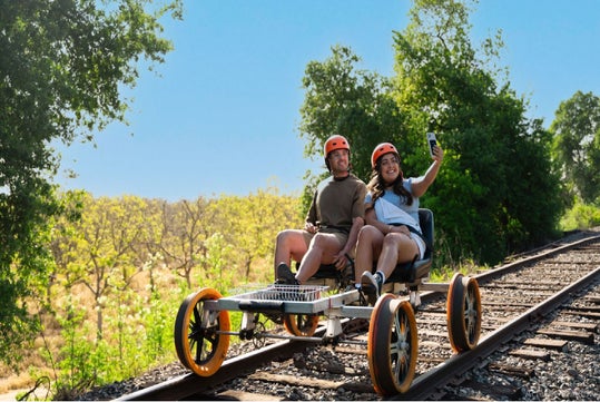 Two people wearing helmets ride a rail cart on train tracks through a scenic landscape, with one person holding a smartphone for a selfie.