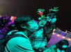 An adult wearing a mask holds a child in a plaid jacket and reindeer antlers as the child raises their arms at an outdoor nighttime event with colorful lights.