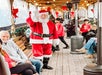 Santa Claus waves while walking down a decorated outdoor corridor, with people, including children in red shirts, seated and standing nearby.