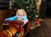 Young girl sits on a wooden toy train in front of a decorated Christmas tree with wrapped presents on a wooden floor.