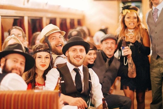 A group of people dressed in vintage attire smiles and poses together at a lively indoor gathering, some holding drinks.