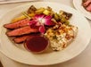 A plate with sliced grilled steak, a serving of cheesy potato gratin, mixed grilled vegetables, a cup of sauce, and an edible flower garnish.