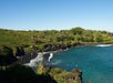 View of a black sand beach with people swimming, surrounded by lush greenery and clear blue water under a clear sky.