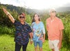 Three people stand outdoors in a grassy field, one pointing to the distance. They appear to be enjoying a sunny day, with hills and trees in the background.