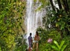 Two people stand on lush green foliage facing a tall waterfall, surrounded by dense tropical vegetation and trees.