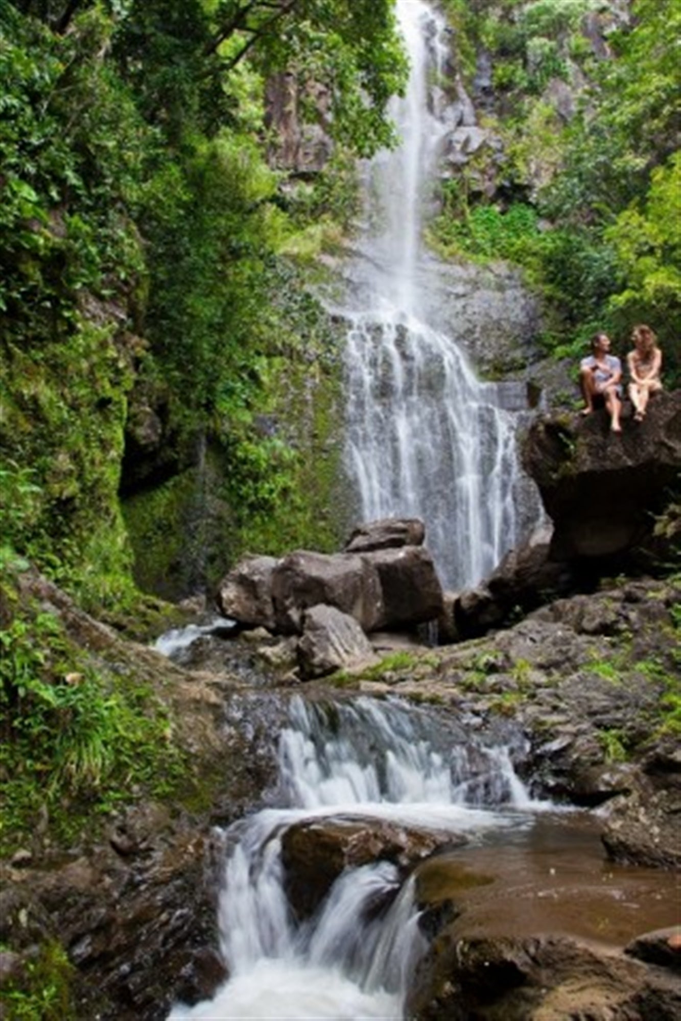 A tall waterfall cascades down rocks surrounded by lush greenery, with two people sitting on a large boulder near the falls.