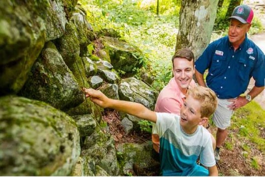 A boy reaches toward a moss-covered rock wall, accompanied by two men, in a forested area.