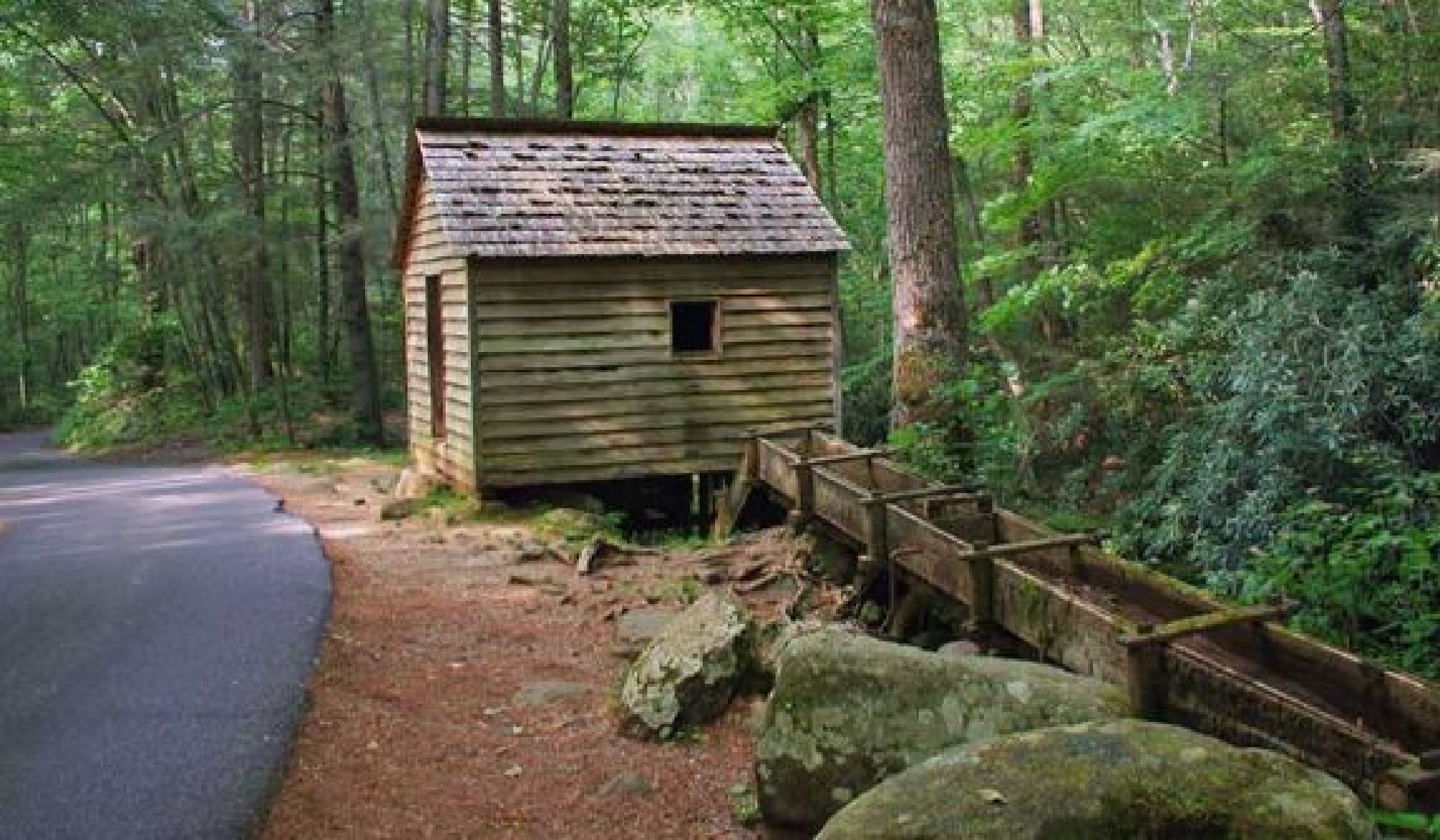 A rustic wooden cabin with a slanted roof and a water chute sits near a paved road, surrounded by dense forest and large rocks.