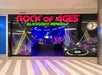 Entrance to "Rock of Ages Blacklight Minigolf" with bright neon lights, colorful graffiti, and musical themes. Bow counter visible inside.
