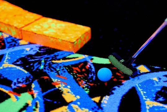 A mini golf putter aims at a bright blue golf ball on a colorful, patterned carpet with neon orange and blue accents.