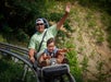 An adult and child ride together on an outdoor roller coaster, both smiling and raising their hands as they move along the track surrounded by greenery.