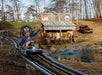 A child rides a mountain coaster past a pond, with a rustic general store and an old car in the background on a grassy hillside.