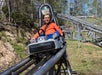 A child in an orange shirt rides a single-car roller coaster outdoors on a sunny day, surrounded by trees and metal tracks.