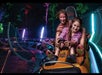 Two people smiling on a brightly lit roller coaster ride with colorful lights in the background.