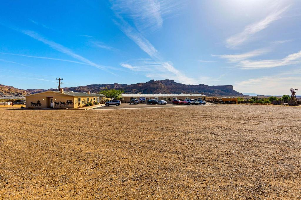 A gravel parking lot with several parked cars in front of a low building. In the background, there are rocky hills under a clear blue sky.
